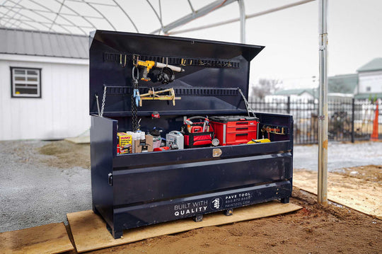 A large, industrial storage container known as Quick-E-BARBoxx, which is black in color, is open and showing various tools and equipment organized inside it. The container is placed outdoors on a wooden palette, and there is a building in the background.